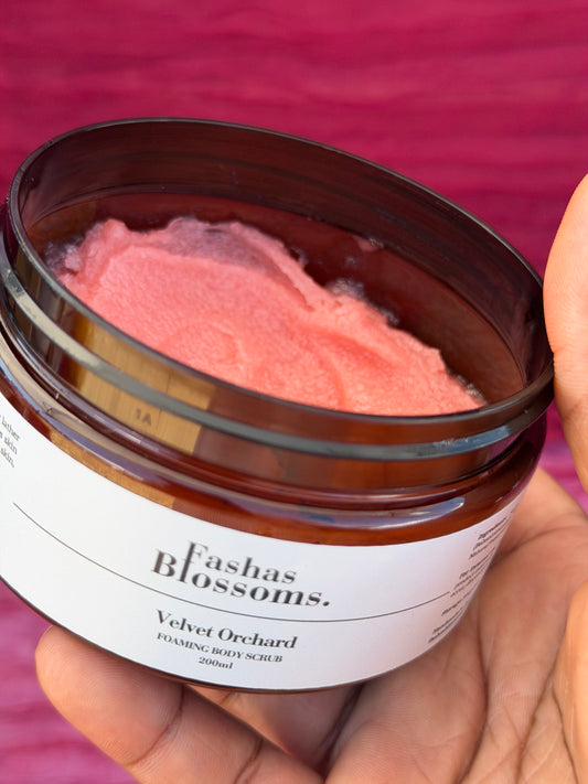 A hand holding an open amber glass jar of Fashas Blossoms Velvet Orchard Foaming Body Scrub, showing the pink scrub inside, set against a textured magenta background.