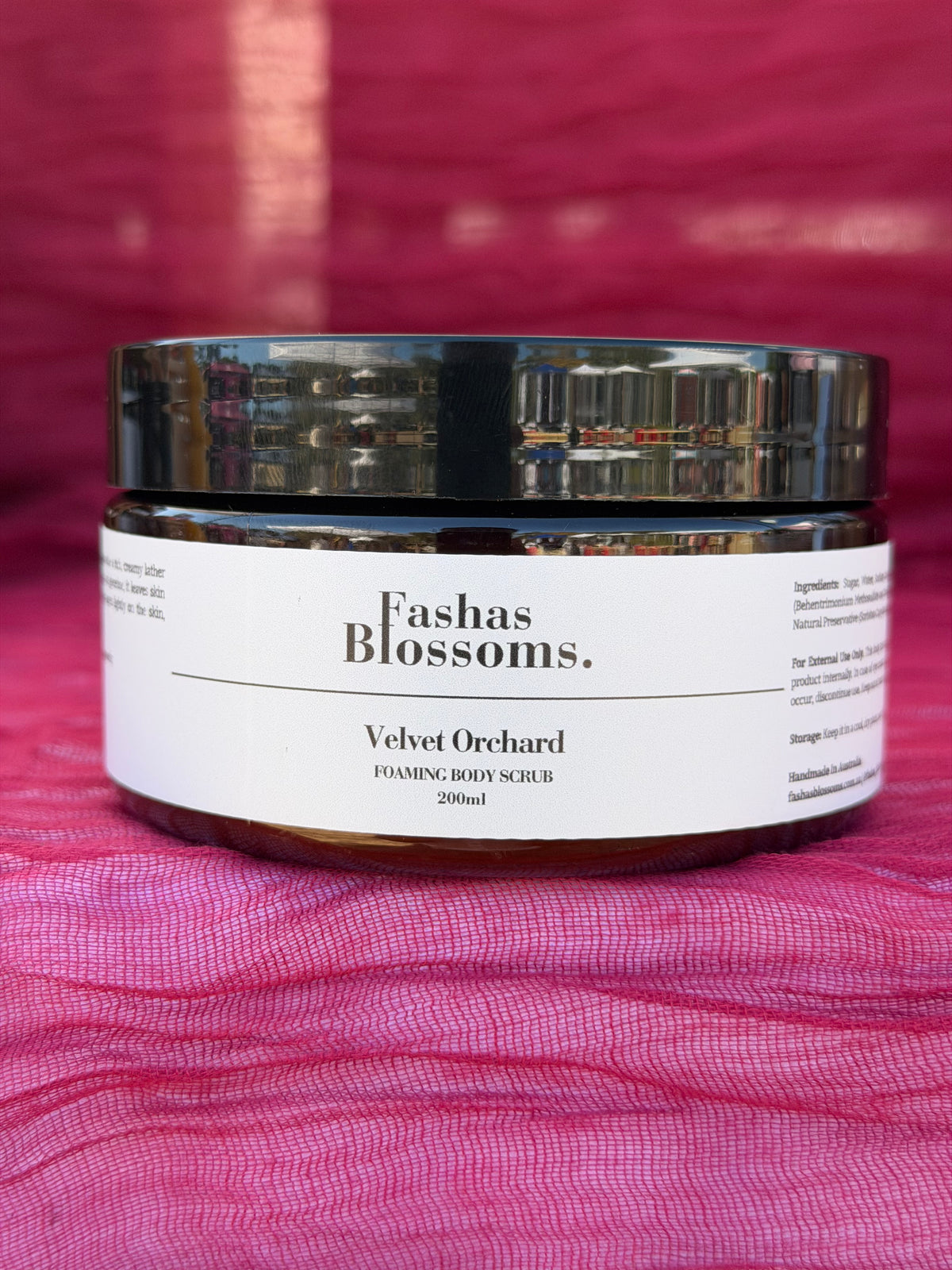 A jar of Fashas Blossoms Velvet Orchard Foaming Body Scrub, featuring a white label and a reflective black lid, set against a textured magenta-pink fabric background.