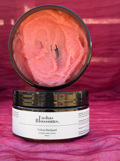 An open jar of Fashas Blossoms Velvet Orchard exfoliating body wash, showing the pinkish-orange scrub inside, with the lid resting on top of the jar against a textured magenta background.