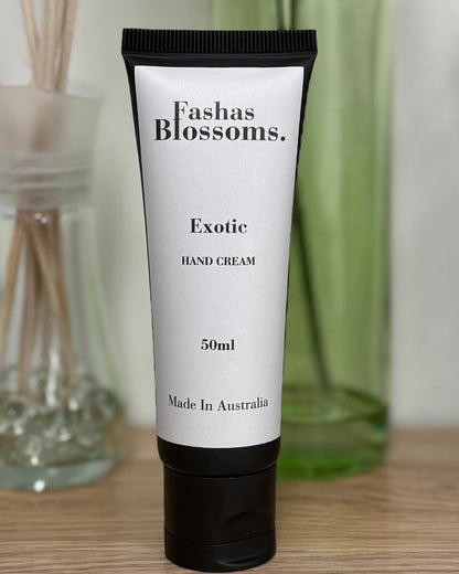 A 50ml tube of Fashin Blossoms Exotic Body Cream stands on a wooden desk, styled with a green glass vase of flowers, a reed diffuser, and a black pencil holder in the background.