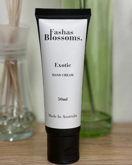 A 50ml tube of Fashin Blossoms Exotic Body Cream stands on a wooden desk, styled with a green glass vase of flowers, a reed diffuser, and a black pencil holder in the background.